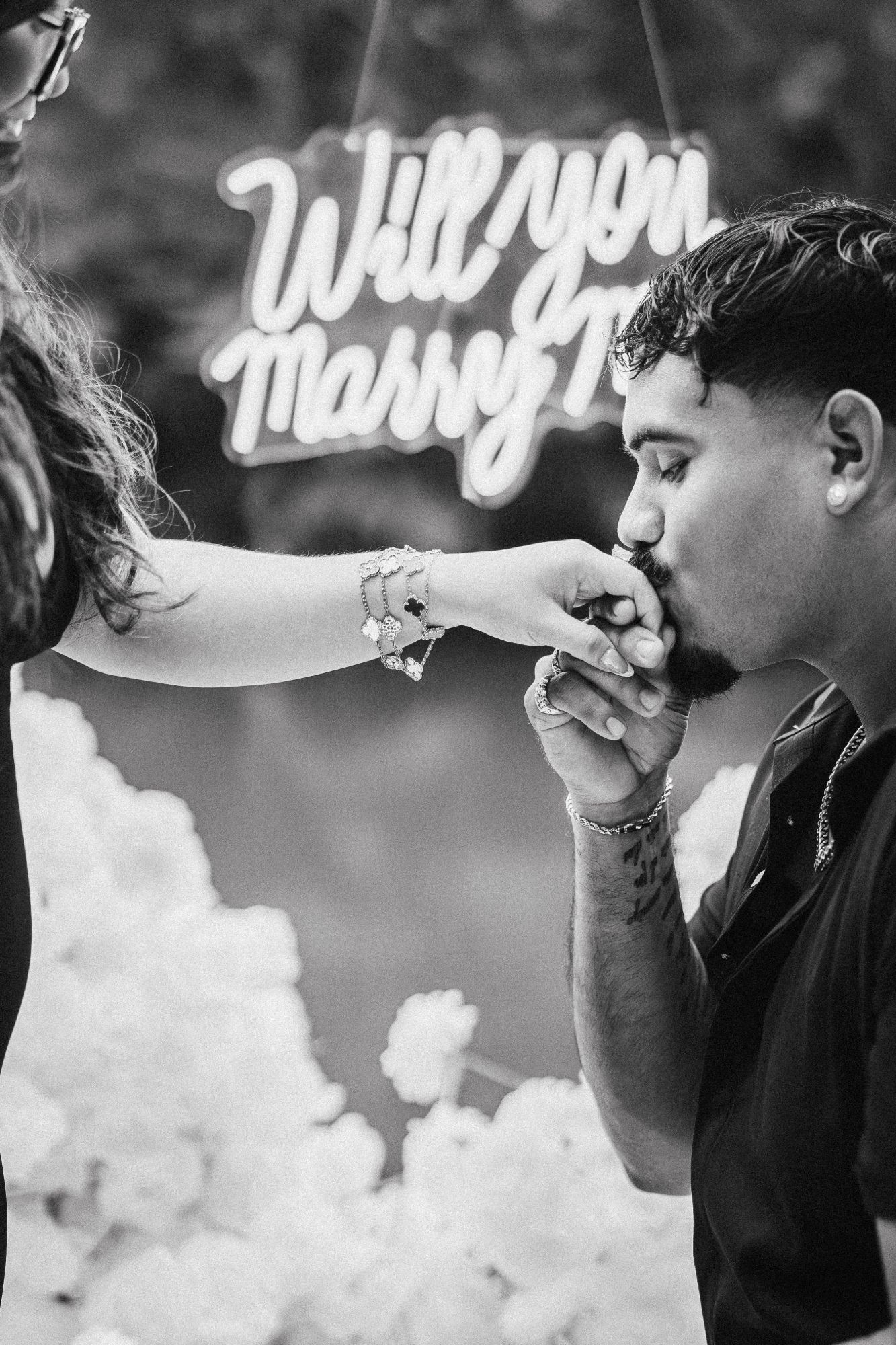 Black and white photo of a hand kiss after the proposal, with a “Will you marry me?” neon sign behind