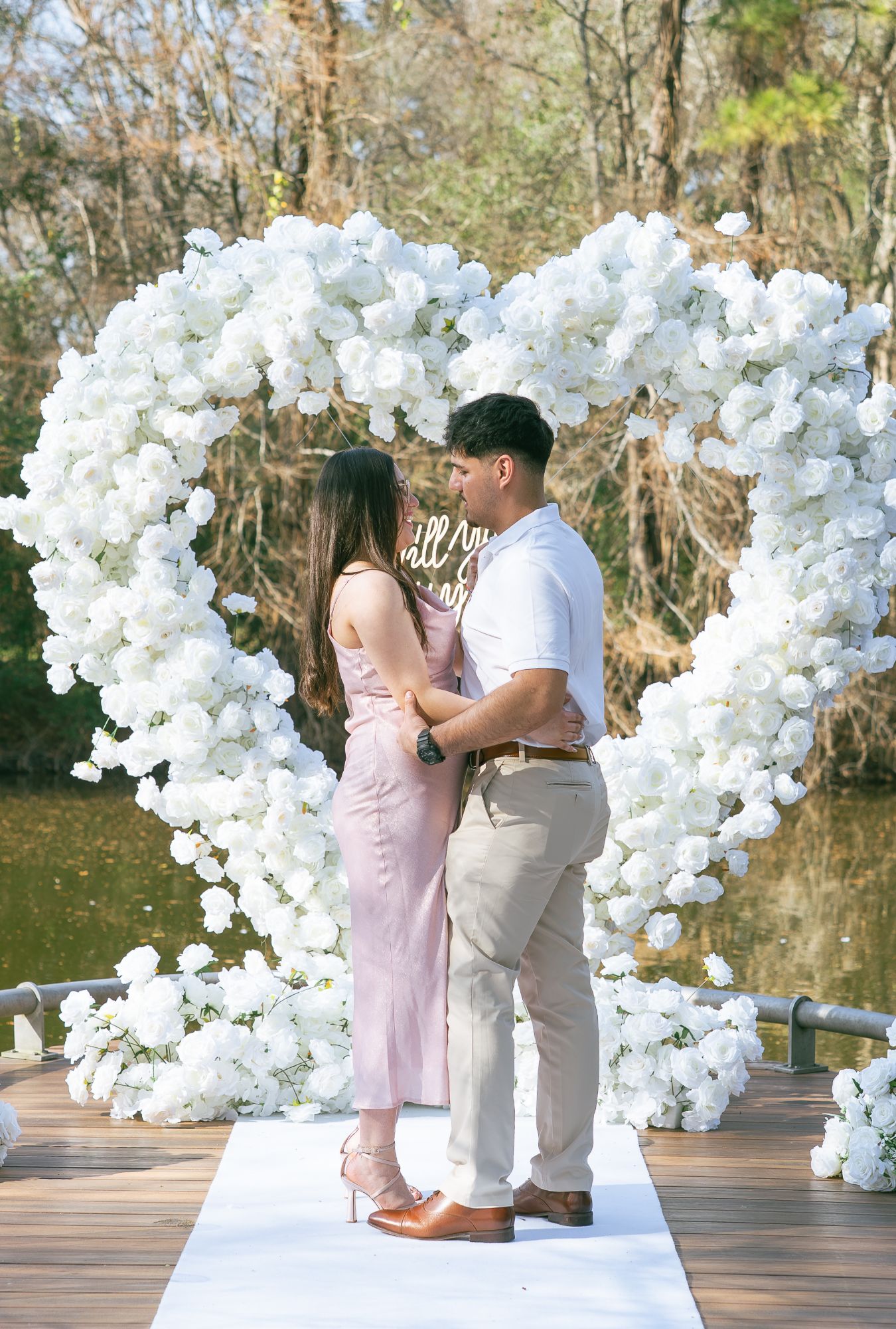 Couple embracing under a white rose heart arch on a lakeside dock after a surprise proposal in Houston