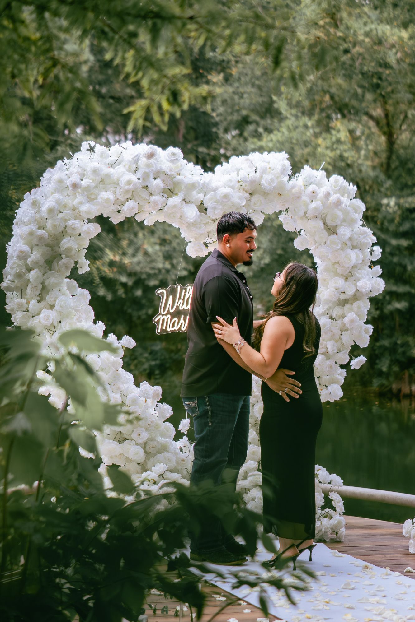 Newly engaged couple embracing in front of a white rose heart arch at a surprise proposal in Houston
