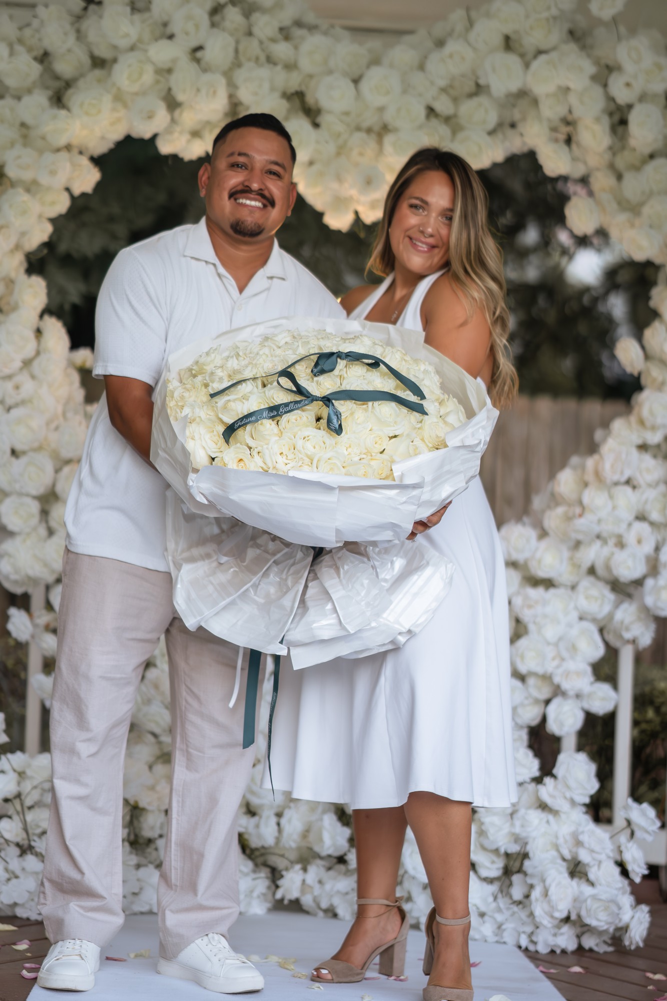 Engaged couple holding a 100 white rose bouquet with a custom ribbon in front of a floral heart arch proposal setup