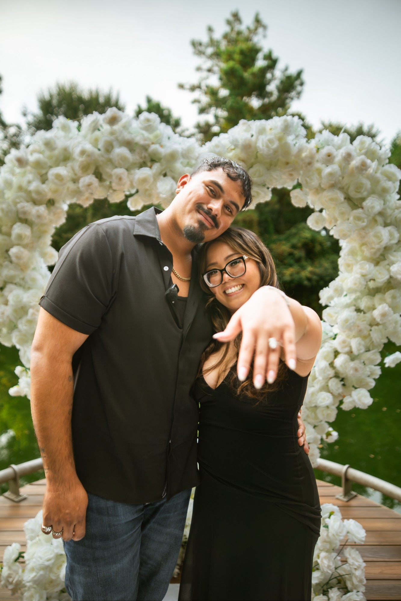 Newly engaged couple showing off the ring in front of a white rose heart arch