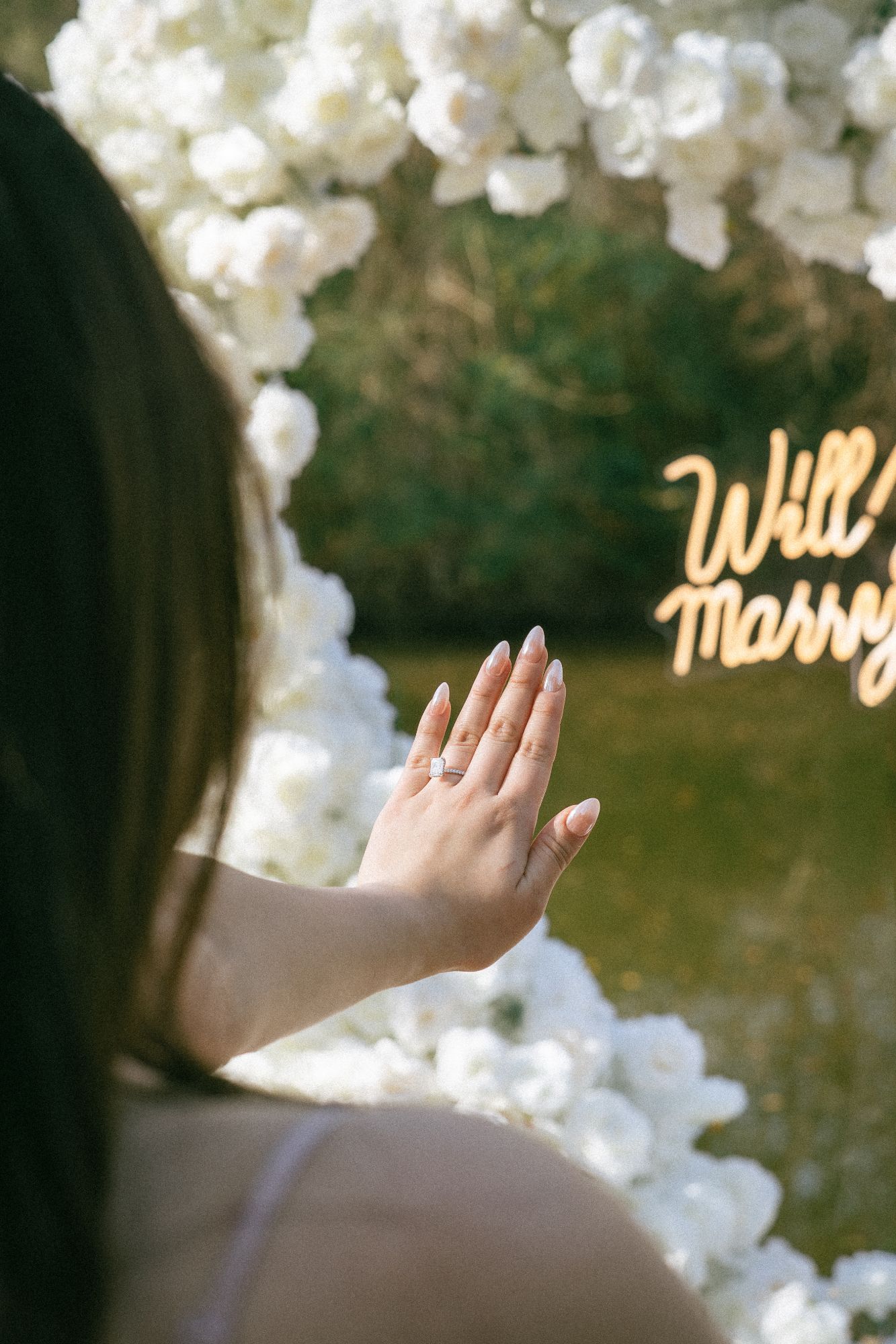 Close-up of an engagement ring with white roses and a glowing “Will you marry me?” sign
