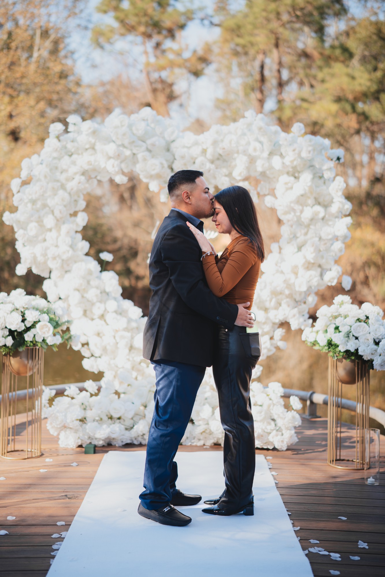 Forehead kiss in front of a white rose heart arch with gold floral pedestals and a white carpet