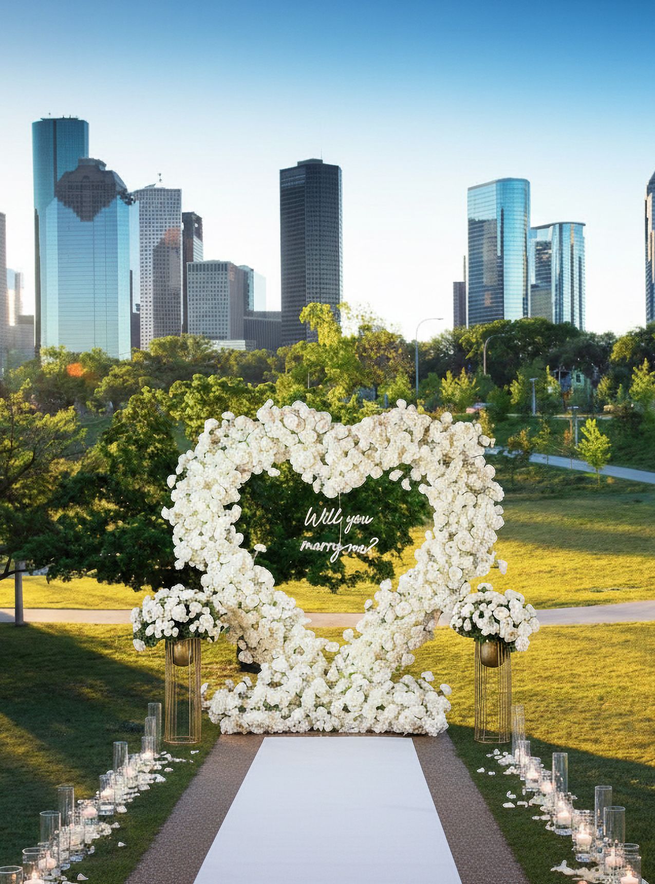 White rose floral heart arch proposal setup with candles, floral pedestals and white carpet in a Houston park with the downtown skyline