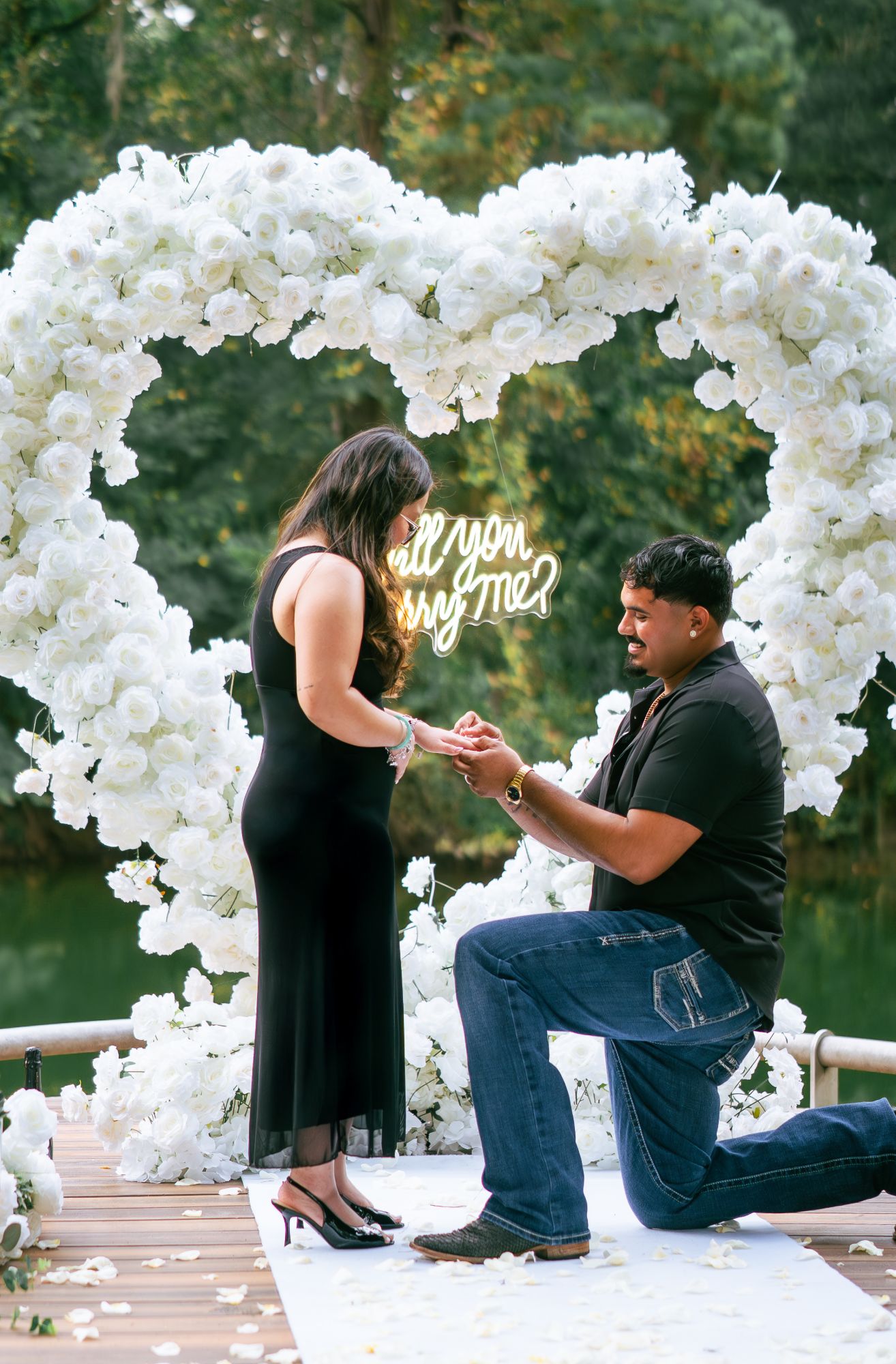 Placing the engagement ring on her finger under a white rose heart arch with a neon “Will you marry me?” sign