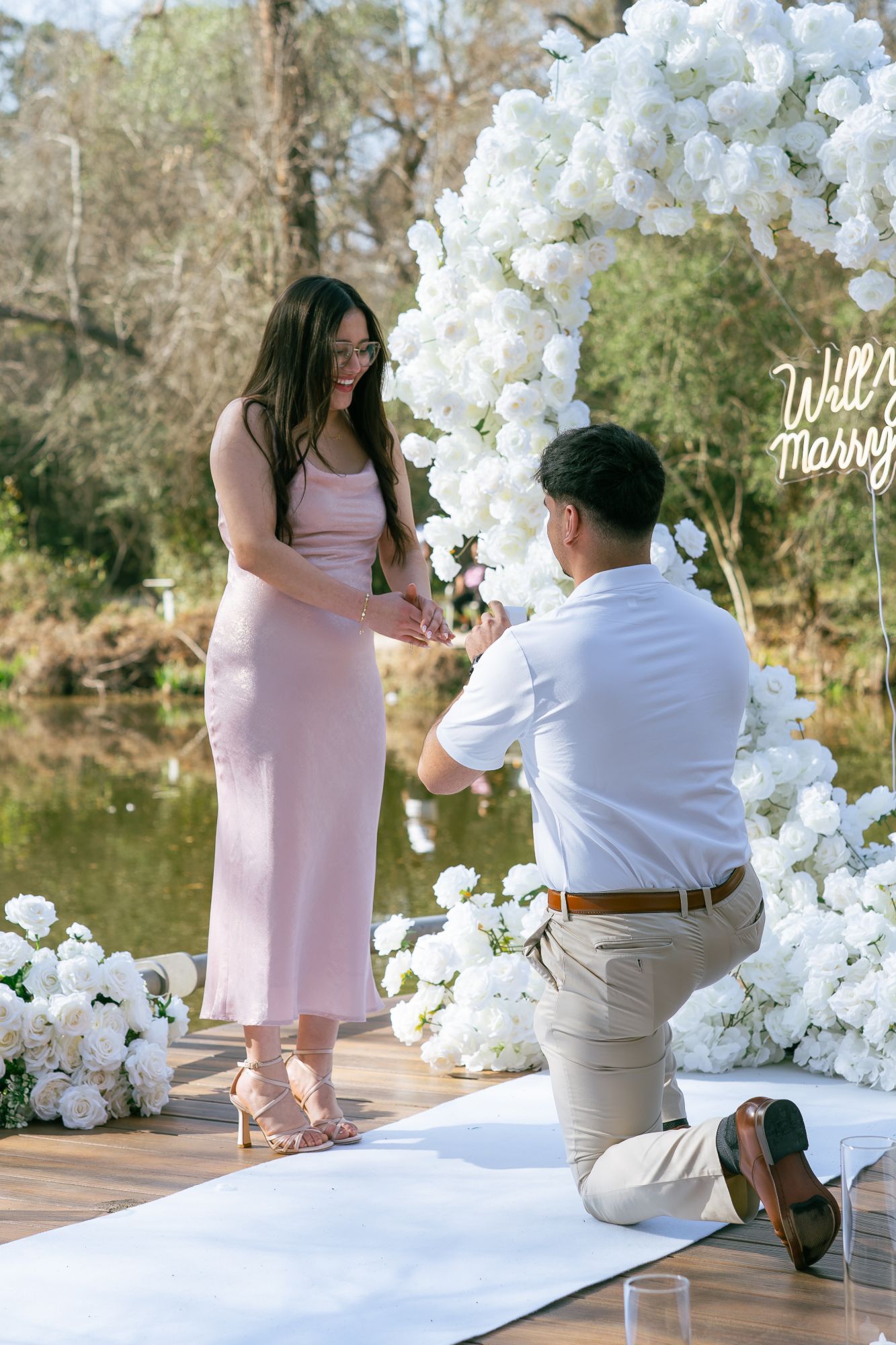 Man on one knee proposing under a white rose heart arch with a “Will you marry me?” neon sign