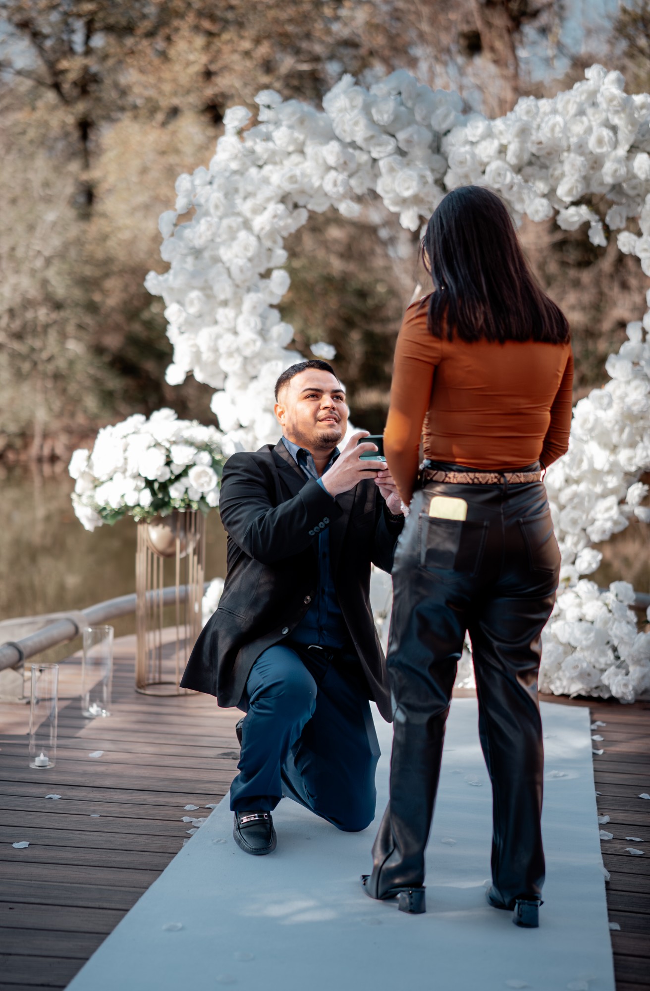 Man kneeling with an open ring box in front of a white rose arch on a dock