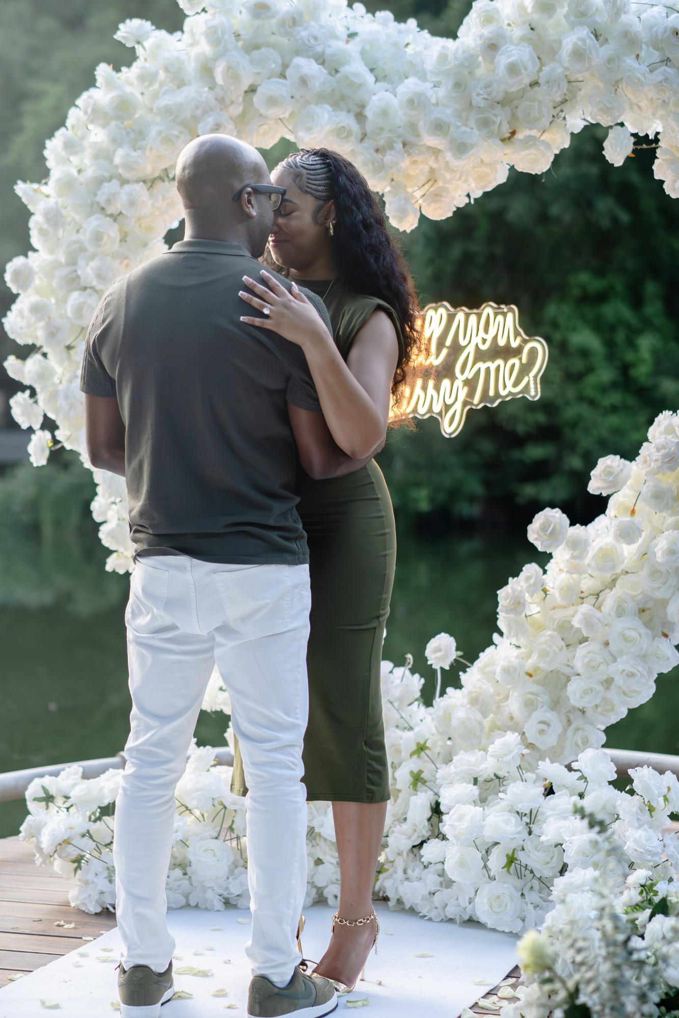 Couple embracing after the proposal under a white floral heart arch with a “Will you marry me?” neon sign